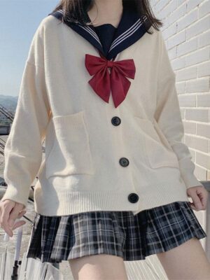Cream white Lolita cardigan with navy sailor collar and red bow tie, front upper-body view on female model against outdoor brick wall background with plaid pleated skirt