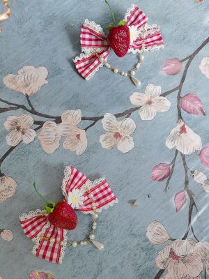 Strawberry gingham red-white checkered Lolita hair bow accessory with pearl drop chains and white lace trim on floral fabric background flat lay top-down view