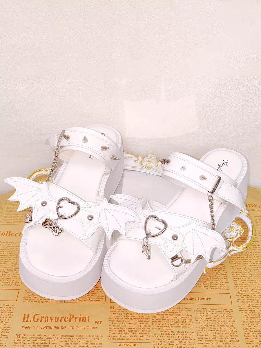 Front overhead view of white Gothic Lolita sandals on newspaper background displaying pristine white patent leather, bat wing charms, heart metal buckles, and delicate chain pendants with open toe design