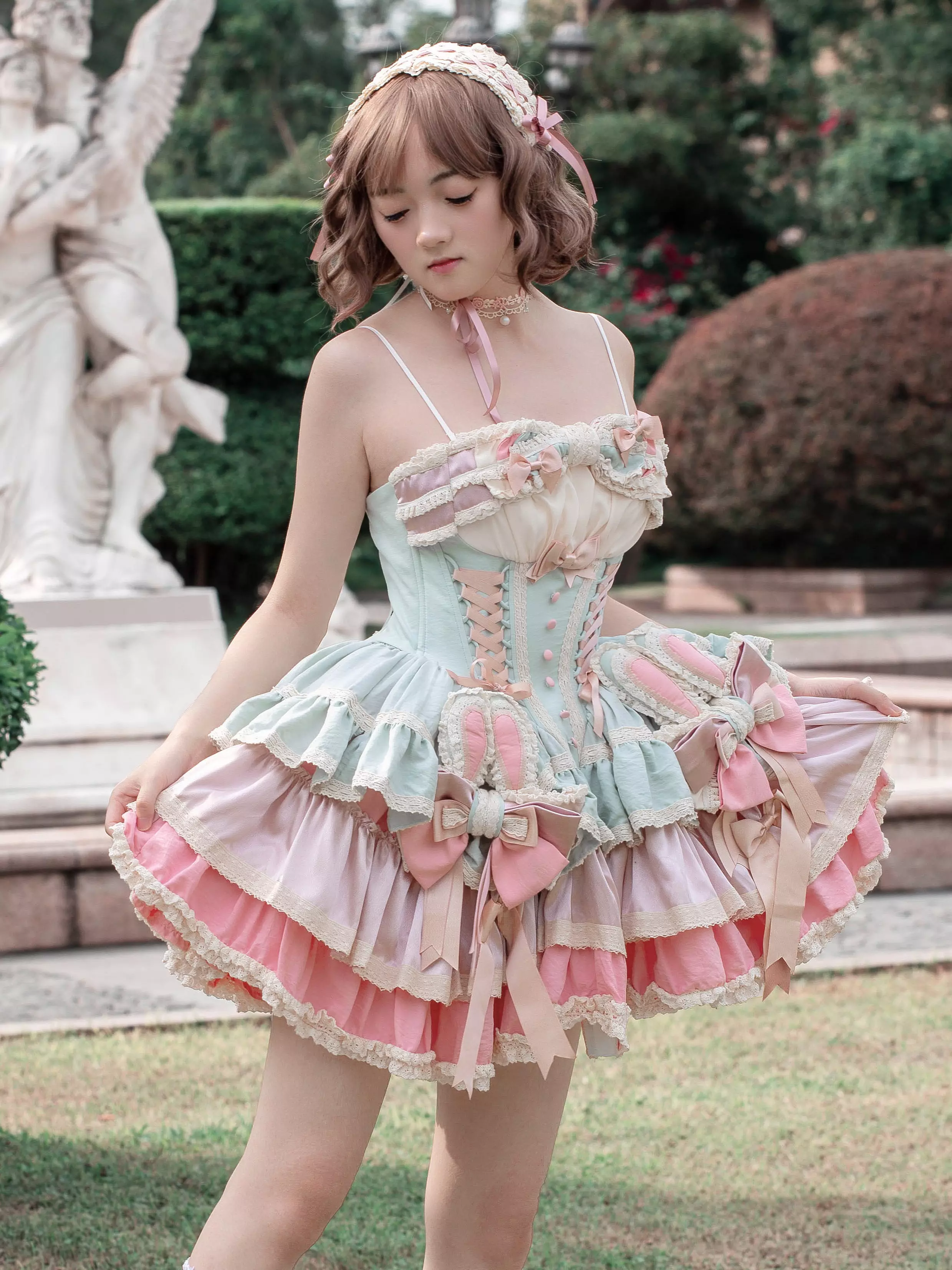 Female model wearing pastel pink sweet lolita bunny ear fairy dress in outdoor garden setting with stone angel statues background