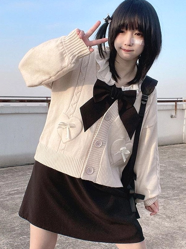Beige lolita cardigan with large black bow tie and heart pockets worn with black skirt in full-length rooftop three-quarter pose