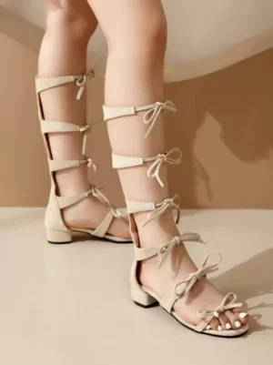 Beige lolita gladiator sandals rear side view, featuring multiple bow embellishments on calf-length tie straps, low block heel, open round toe design, suede matte finish on female model legs