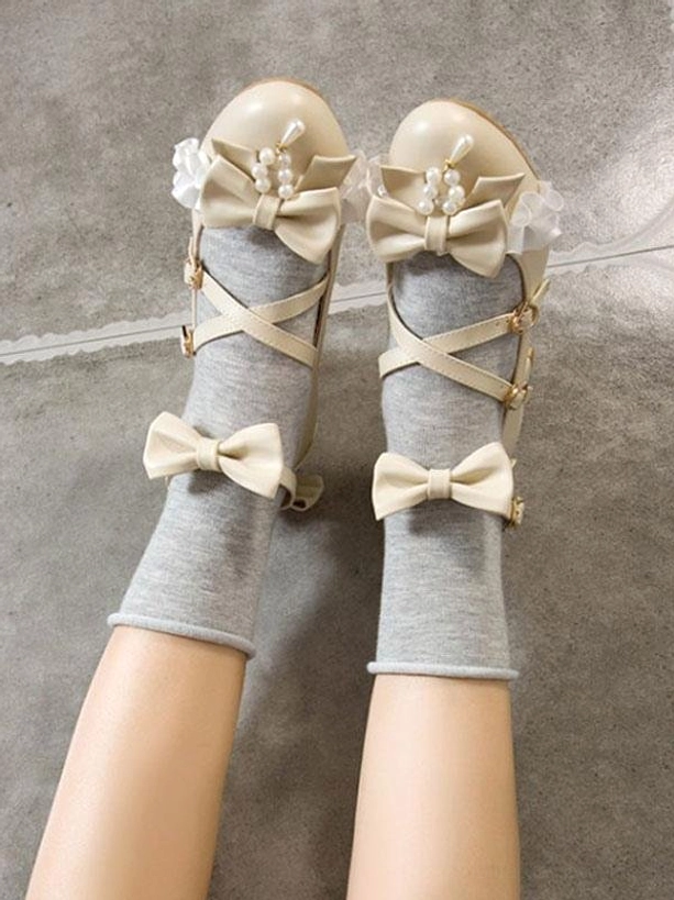 Beige lolita chunky heel pumps with cross ankle straps and dual bow decorations plus pearl embellishments shown in overhead view of model's legs wearing gray crew socks, displaying top-down perspective of shoe construction