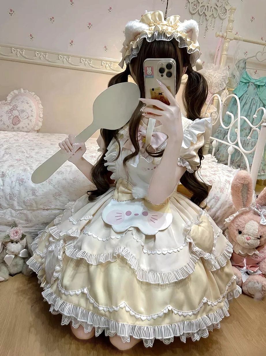 Beige sweet lolita cat dress front seated view with ruffled layered skirt, cat face apron, plush toy bedroom background, model with crown bear ear headpiece holding mirror