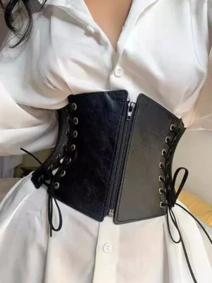 Close-up waist-level view of black corset belt worn with white long sleeve shirt, showcasing matte faux leather texture and adjustable side lace-up bows
