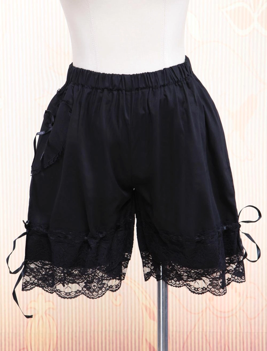 Black cotton lolita bloomers front three-quarter view showing elastic waist, delicate lace hem trim, and side ribbon accents on dress form with patterned backdrop