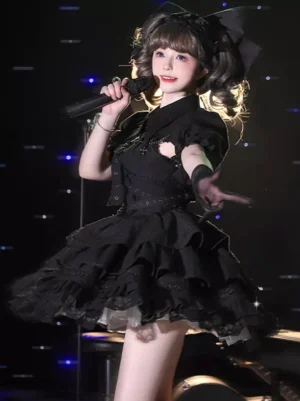 Black gothic lolita jumper dress with tiered ruffles and chain cross accents worn by female model performing on stage with microphone