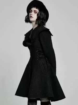 Three-quarter side front angle of black gothic lolita overcoat showing decorative pom-pom on chest, ruffled collar detail, waist lacing, structured silhouette with fedora styling