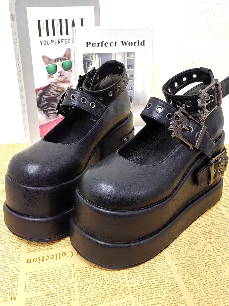 Pair of black Gothic Lolita pumps front three-quarter view on fashion magazine backdrop showing smooth black PU leather skull buckle decorations and wedge platform sole