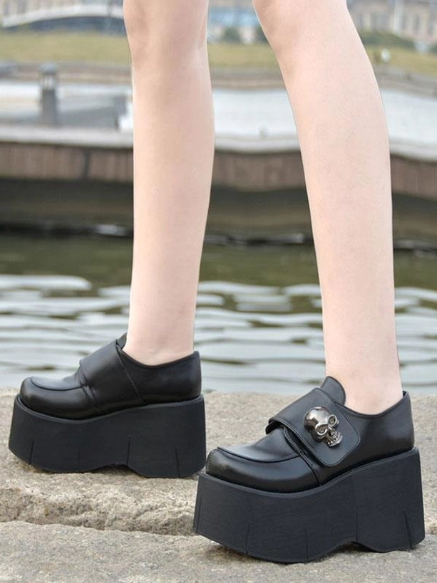 Black gothic skull platform lolita pumps side-view worn outdoors showing glossy pu leather finish ultra-thick flatform sole and polished skull buckle detail on stone ground waterfront setting