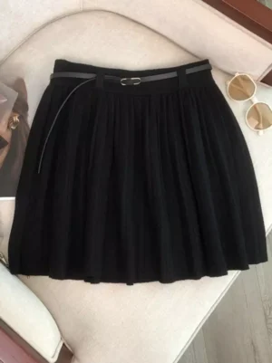 Black knitted pleated mini lolita skirt with high waist A-line silhouette and leather waist belt displayed on sofa with sunglasses