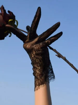 Single gloved hand holding dried dark rose displaying black lace gloves with fringed wrist edge and gothic aesthetic from side upward view, romantic Lolita accessory photography with blue sky backdrop