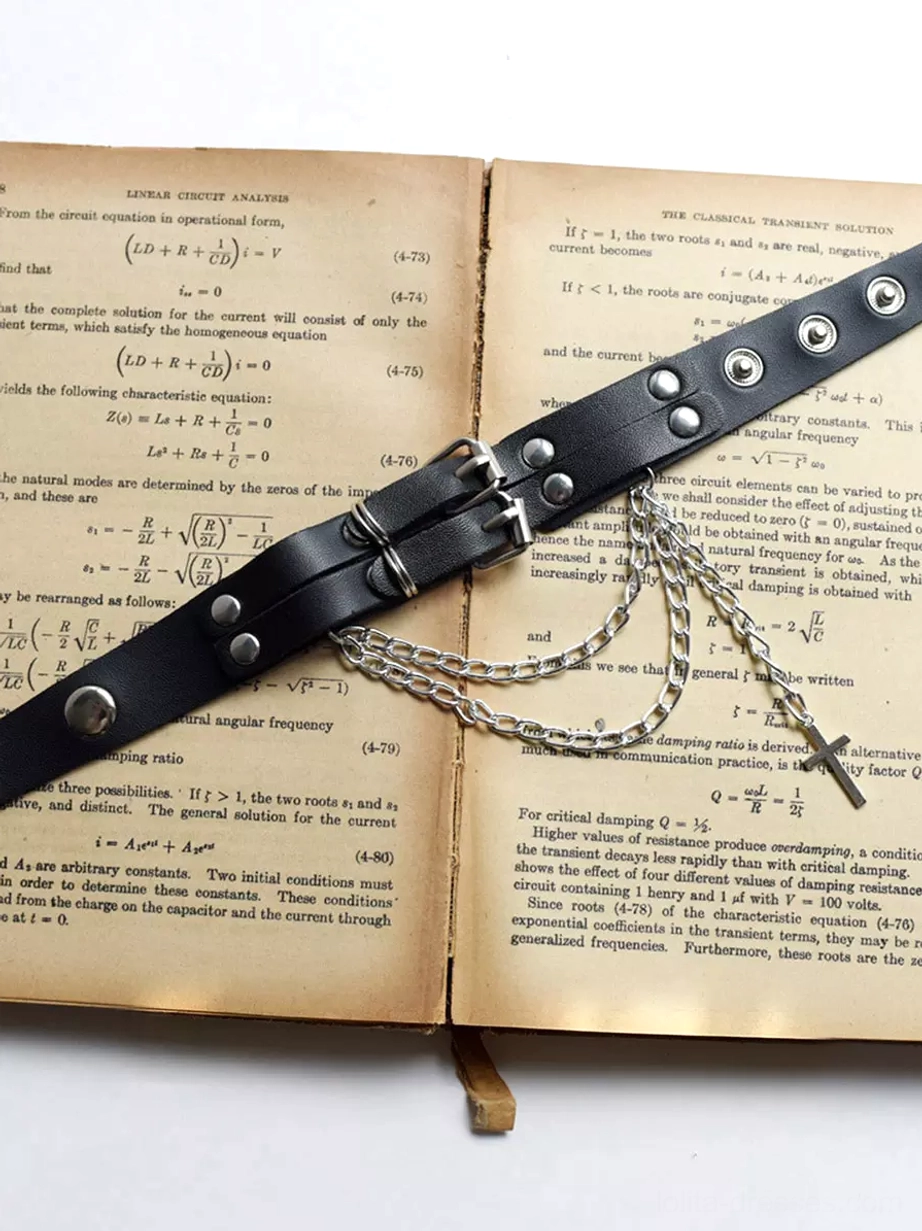 Black leather gothic arm band flat lay top-down view on vintage English textbook showing silver hardware, rivets, chains and cross pendant, product detail shot