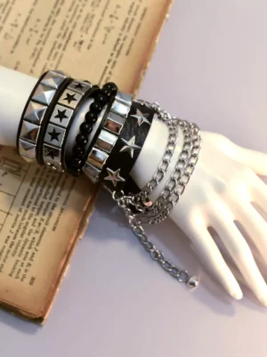 Gothic punk multi-layer black leather bracelet with silver star studs and dangling metal chains displayed on white mannequin hand with vintage book backdrop, showing side overhead view of stacked rivet bracelets