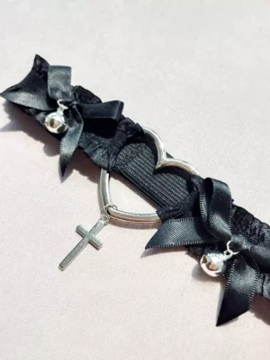 close up detail view of black thigh garter showing adjustable metal buckles ruffled elastic strap and silver heart cross charm with bell