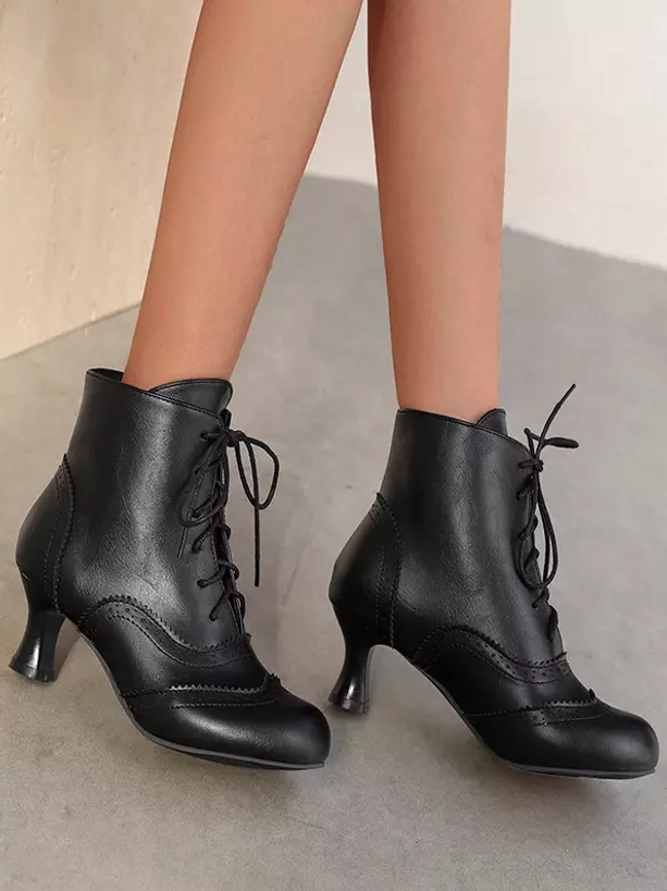 Black vintage ankle boots in frontal flat view on female lower legs showing lace-up closure, sleek PU leather finish, and timeless silhouette against plain light gray background
