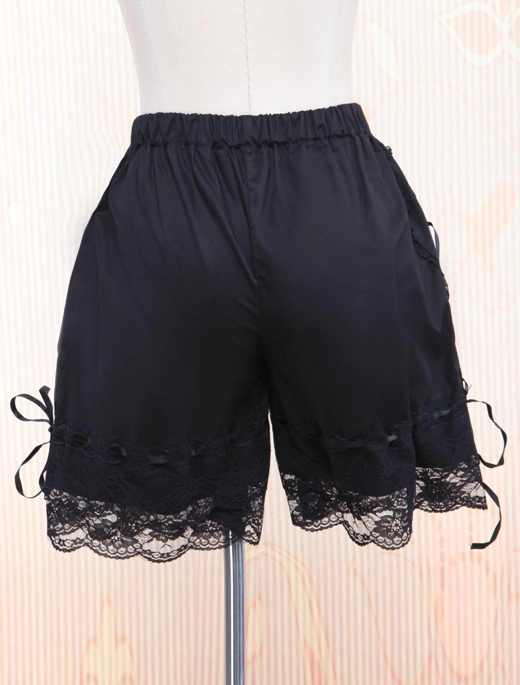 Black lolita bloomers rear back view showing elastic waistband, bilateral lace hems, and leg-side ribbon bows on dress form with striped background
