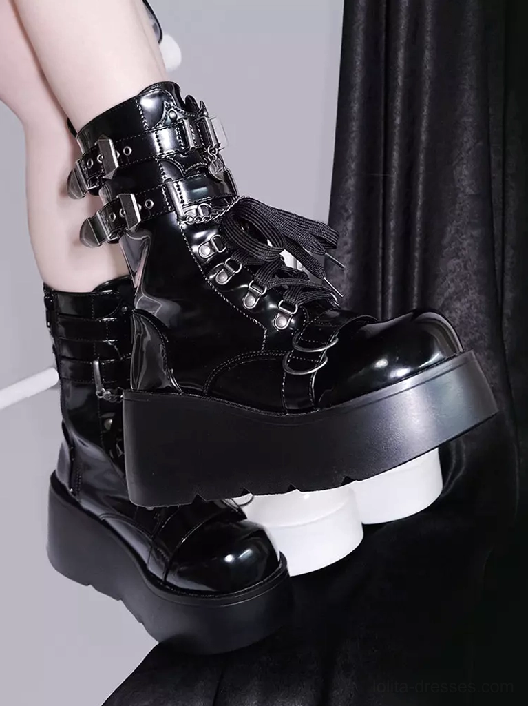 Close-up side view of black platform ankle boots with metal buckles fringe trim and glossy chunky platform sole against dark fabric backdrop