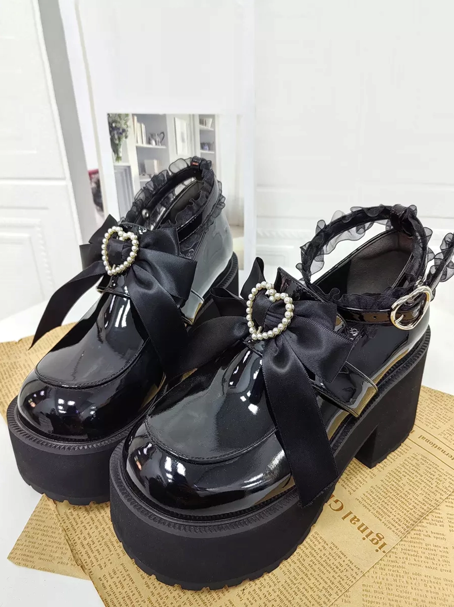 Front three-quarter view of black gothic lolita platform pumps pair displayed in indoor setting with newspaper and door background, showing chunky platform block heel and heart rhinestone buckle details