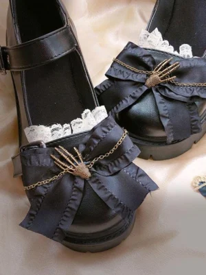 Pair of black Lolita platform shoes flat lay with metal chain skeleton hand bow, lace frill trim, and adjustable buckle ankle strap on satin background
