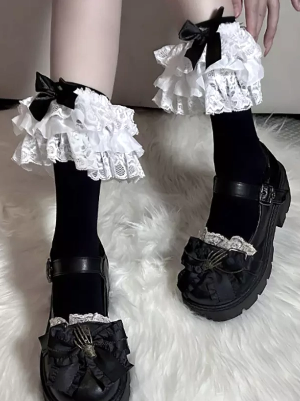Black lolita ruffle ankle socks with prominent lace frill and satin bow worn with matching black platform mary jane shoes, angled close-up view showcasing detailed lace construction and coordinated footwear pairing