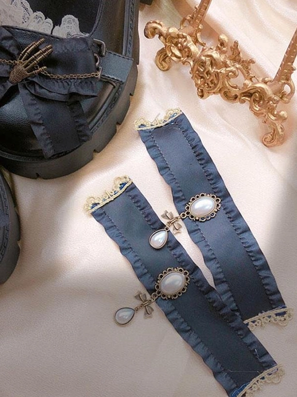 Black Lolita shoes and matching pearl-decorated ankle accessories flat lay on satin background with golden decor, showcasing skeleton hand bow and ruffled edge design