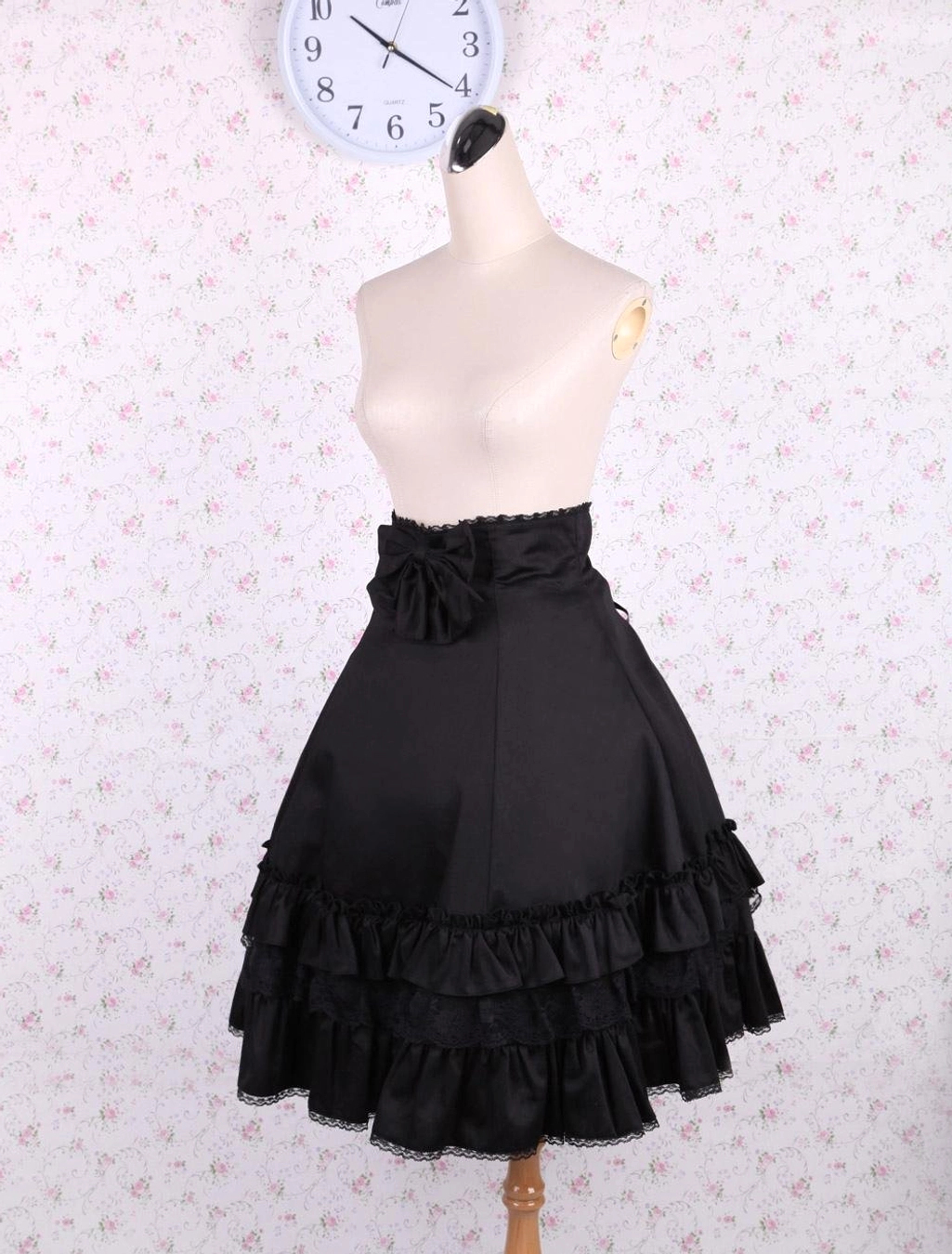 Black high-waisted lolita skirt three-quarter angle showing front bow and cascading ruffled lace layers on dress mannequin