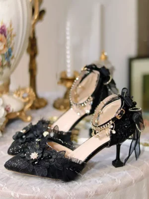 Gothic Lolita black stiletto heels featuring pointed toe with crystal ankle strap and tulle bow heel trim, photographed on vintage porcelain table setting, highlighting dimensional flower embellishments and sparkle fabric upper