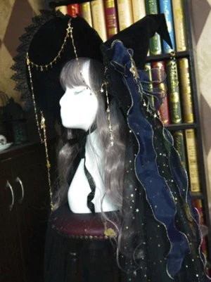 Black lolita witch hat with navy blue mesh bow and gold face chain, left side view on mannequin with grey-brown wig in front of vintage bookshelf background