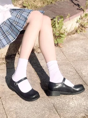 Pair of black Mary Jane Lolita shoes displayed side view, worn on both feet in outdoor pavement setting, showing the solid matte black finish, low block heel, closed round toe, and classic Lolita silhouette with white socks and plaid skirt