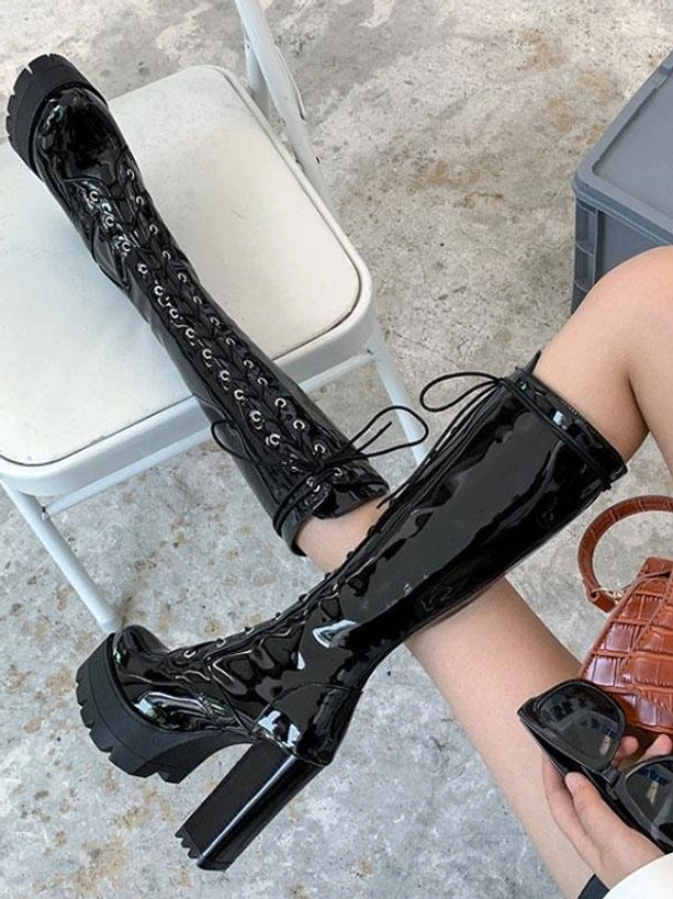 Black glossy mid-calf lace-up boots worn by model on white chair outdoors, showing tall shaft and chunky platform heel from overhead side angle