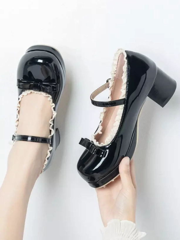 Black patent leather lolita pumps top-down hand-held view showing front bow ruffled collar ankle strap and block heel plain white background
