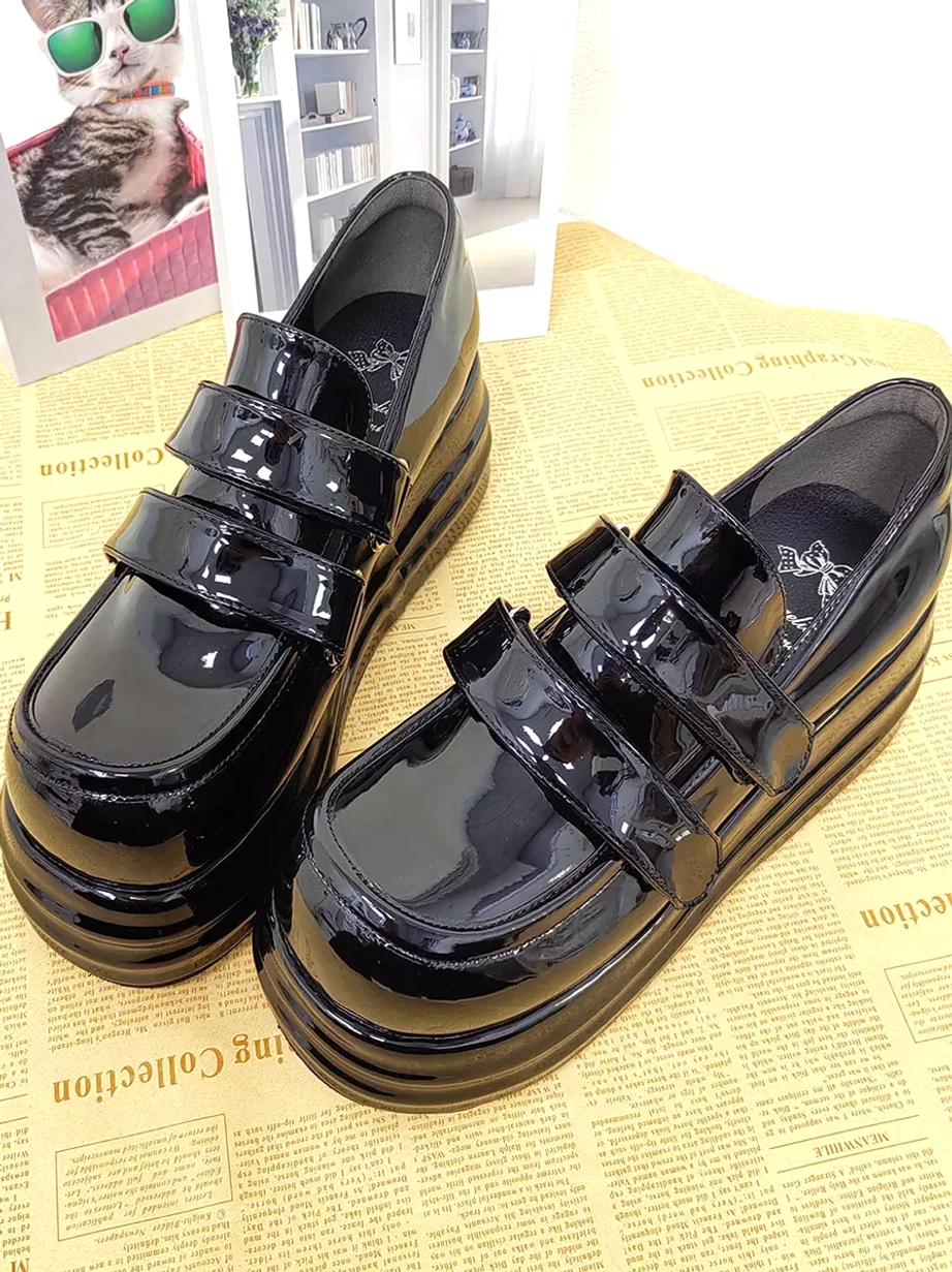 Pair of black glossy patent leather Gothic Lolita platform shoes front oblique top view with double velcro straps and decorative newspaper cat themed background