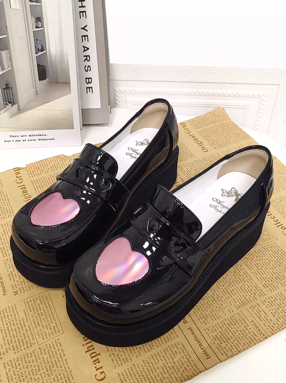 Sweet Lolita black patent leather platform pumps with newspaper prop background, oblique view displaying branded insole and holographic heart detail