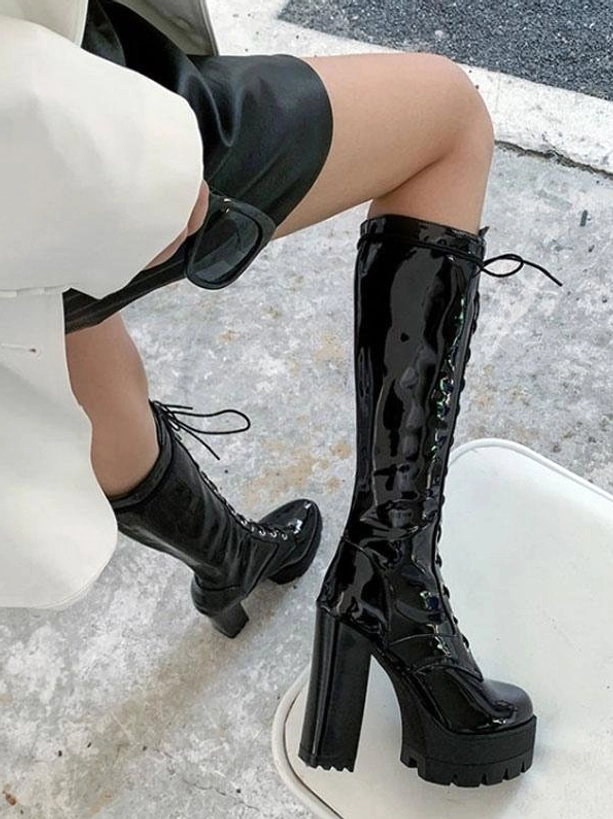 Black patent leather mid-calf boot placed on white chair low side view, displaying block platform sole and high chunky heel construction detail
