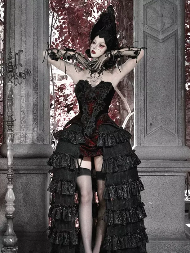 Gothic lolita skirt gown featuring layered ruffled hem and strapless corset with thigh bands, full front shooting angle with model wearing pointed witch hat and stone pillar terrace setting