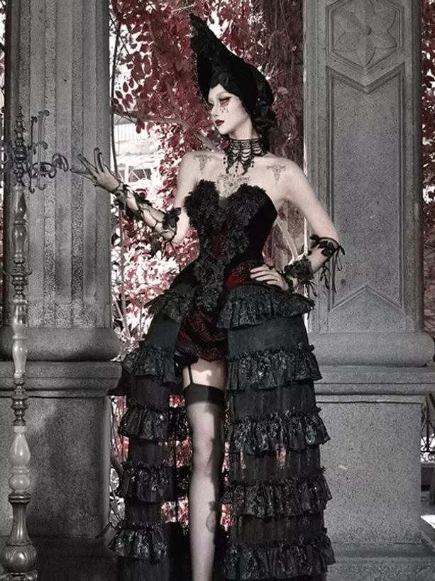 Black and red gothic lolita skirt gown with thigh bands and high-low ruffled hem design, half front view showing model wearing pointed witch hat at stone pillar terrace location