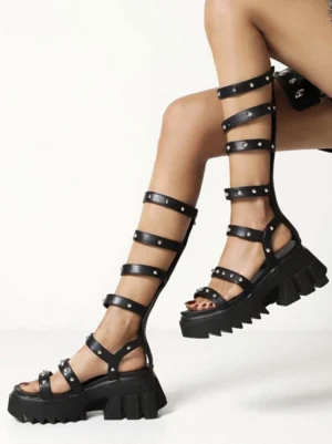 Black gladiator platform sandals with silver rivets, side front view showing multi-strap construction and chunky lug sole, worn on model's leg against white studio background