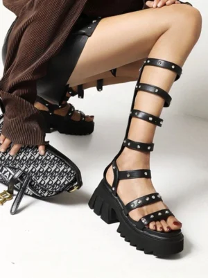 Black rivet gladiator sandals low side angle view displaying calf-length straps with silver cone-shaped rivets and thick platform sole, styled with monogrammed handbag