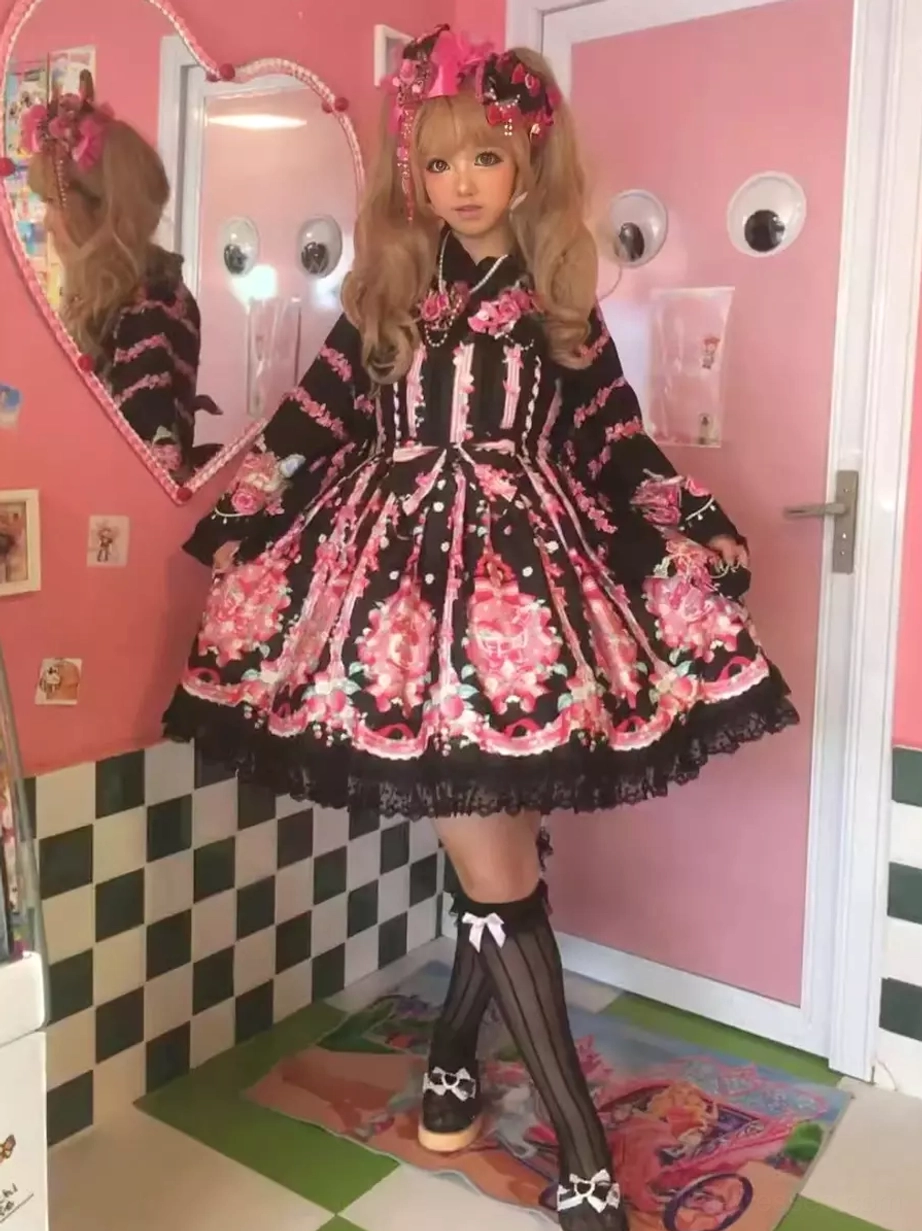Black sleeveless sweet lolita dress with pink strawberry floral print and waist bowknot, worn by cosplay model in retro pink interior setting, showcasing full-body front view with ruffled lace hem and lace collar details