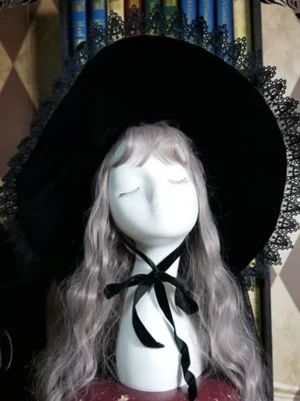Black velvet lolita witch hat with lace trim displayed on mannequin with ash curly wig front flat view against vintage bookshelf backdrop