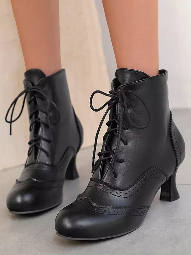 Black vintage lace-up ankle boots with brogue trim and black tapered heel shown from side and frontal-side angle on light gray floor, representing Gothic Lolita aesthetic with classic proportions
