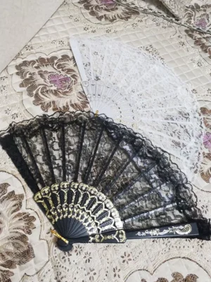 Black and white lace folding hand fan lolita accessory with ornate carved plastic frame and ruffled lace edge displayed on floral quilted fabric background overhead view