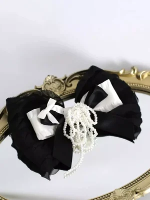 Black and white ruffled lolita bow hair clip with pearl tassel displayed on vintage gold mirror tray, flat lay front view showing intricate fabric details and romantic design elements