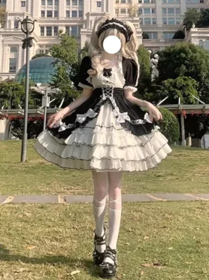 Front full-body view of black and white sweet lolita JSK dress with multi-layer ruffles and front lace-up bodice worn by blonde model on outdoor lawn