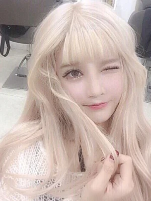 Long blonde wavy synthetic Lolita wig with blunt bangs, front close-up shot showcasing pale blonde curly hair texture and fringe detail, playful styling with wink expression