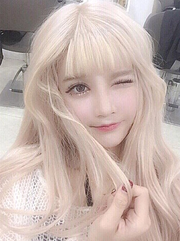 Heat-resistant blonde lolita wig with blunt air bangs and flowing wavy hair on model in hair salon setting, winking pose, showing pale blonde fiber quality and sweet lolita romantic style