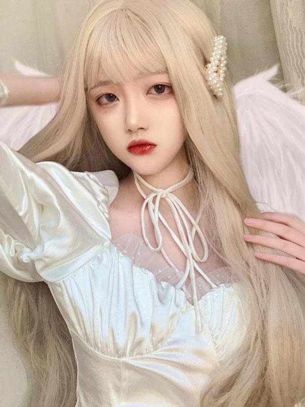 Front view of long blonde Lolita wig with blunt bangs worn by female model in white Lolita dress with angel wing backdrop and pearl hair clip