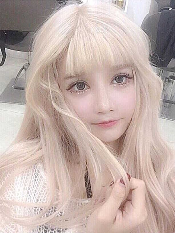 Long blonde lolita wig featuring soft pale blonde wavy hair with neat air bangs on model in salon background holding hair strand, showcasing heat-resistant synthetic fiber texture and sweet lolita aesthetic