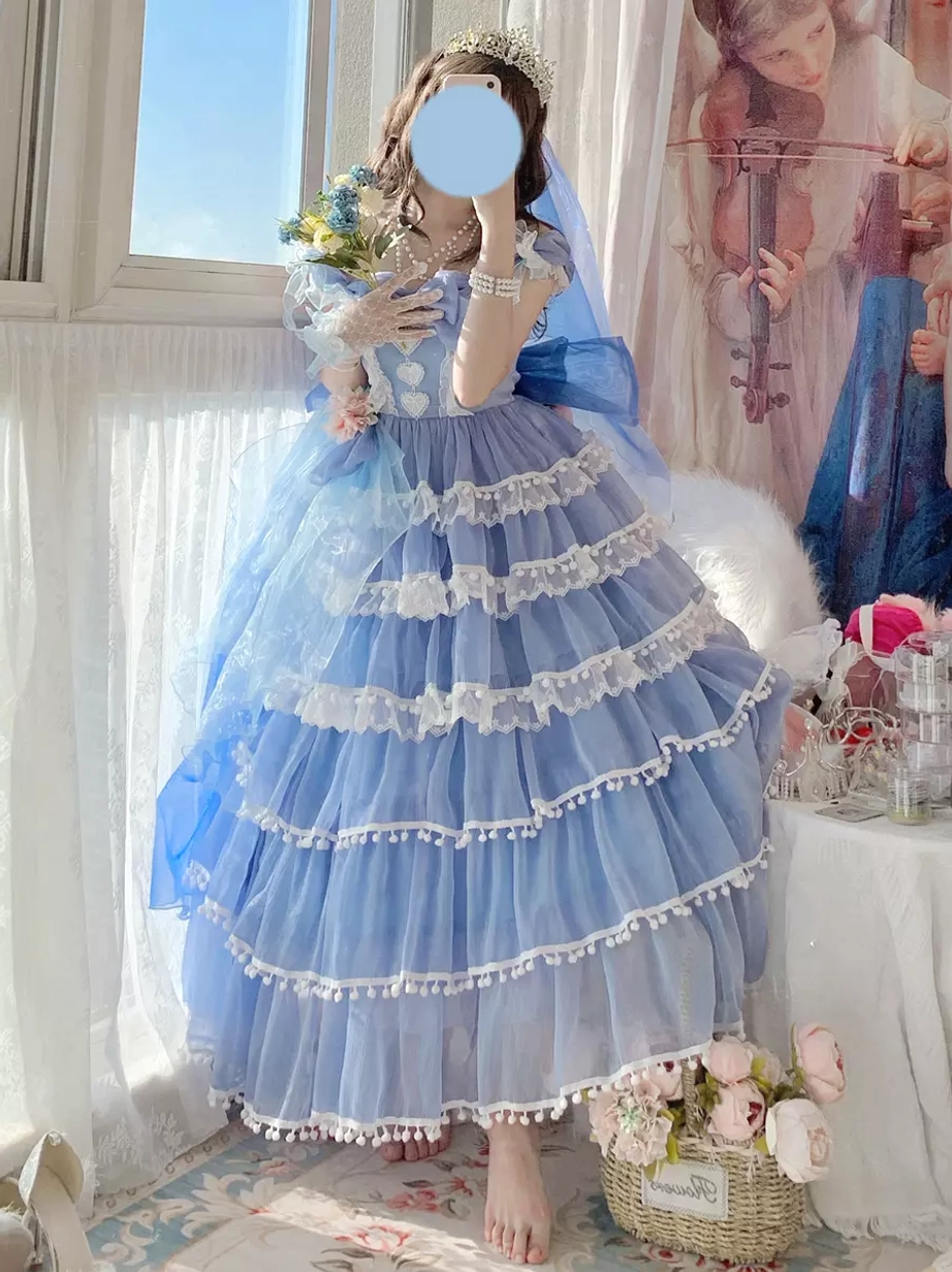 Blue tiered lolita wedding dress with layered tulle skirt, white pom-pom trims, and off-shoulder design featuring tiara and bridal veil